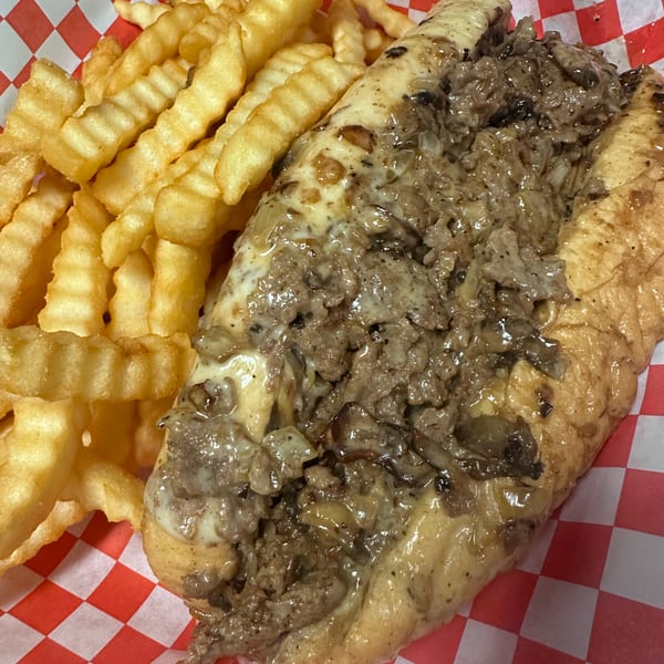 Cheesesteak sandwich with melted cheese and onions, served with crinkle-cut fries in a red and white checkered basket.