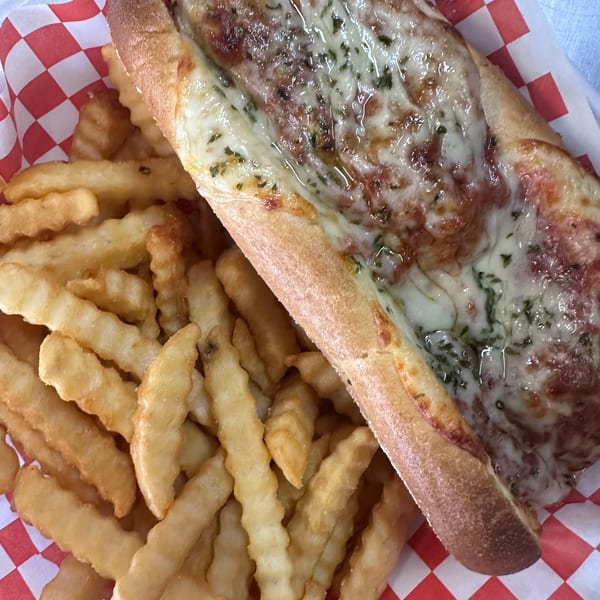 Meatball sub with melted cheese and herbs, served with crinkle-cut fries in a red and white checkered basket.