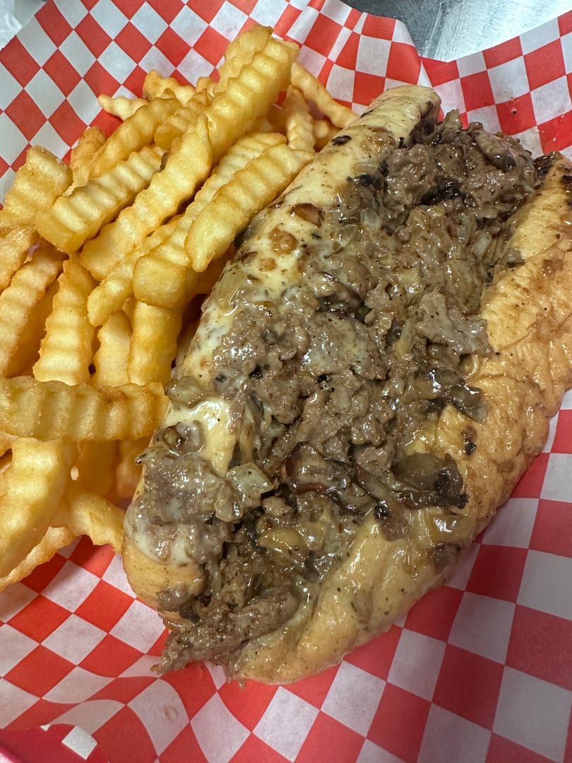 Cheesesteak sandwich with melted cheese and onions, served with crinkle-cut fries on red and white checkered paper.