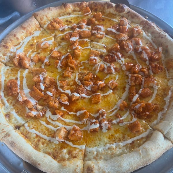 A buffalo chicken pizza with ranch drizzle on a metal tray, with a pizza server underneath.