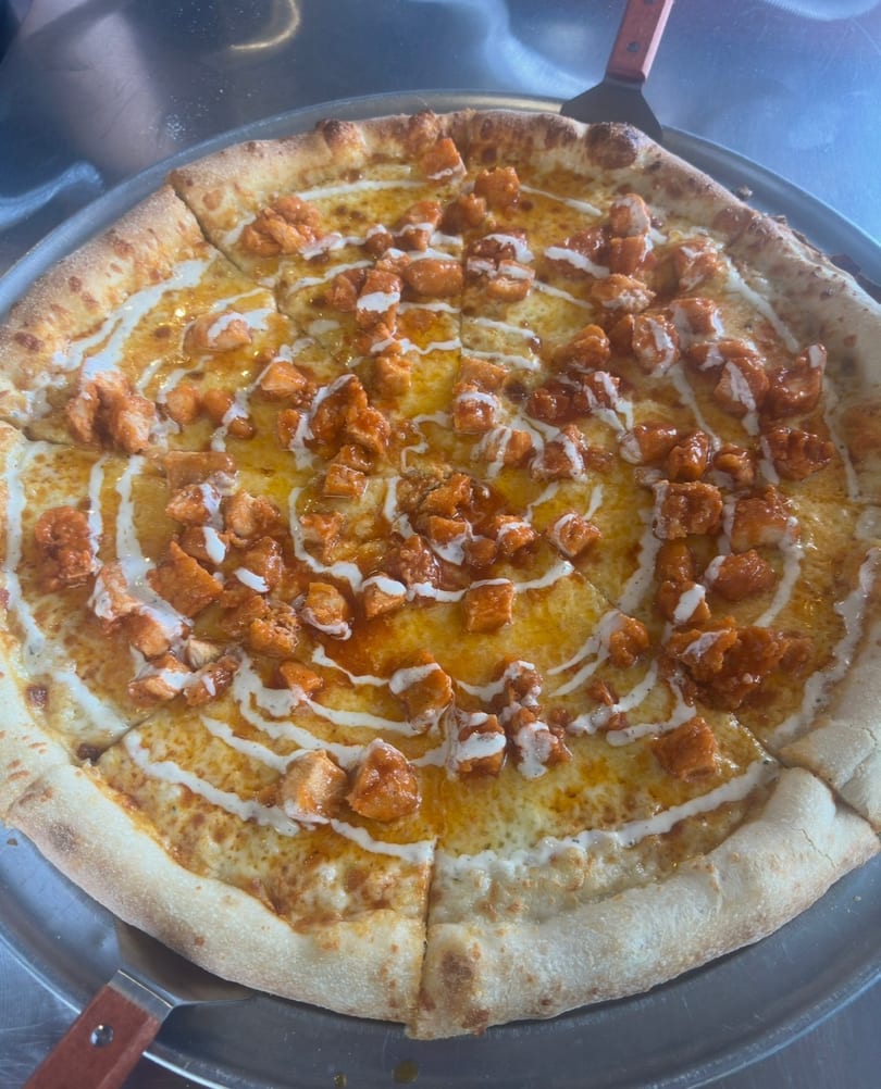 A buffalo chicken pizza with ranch drizzle on a metal tray, with a pizza server underneath.