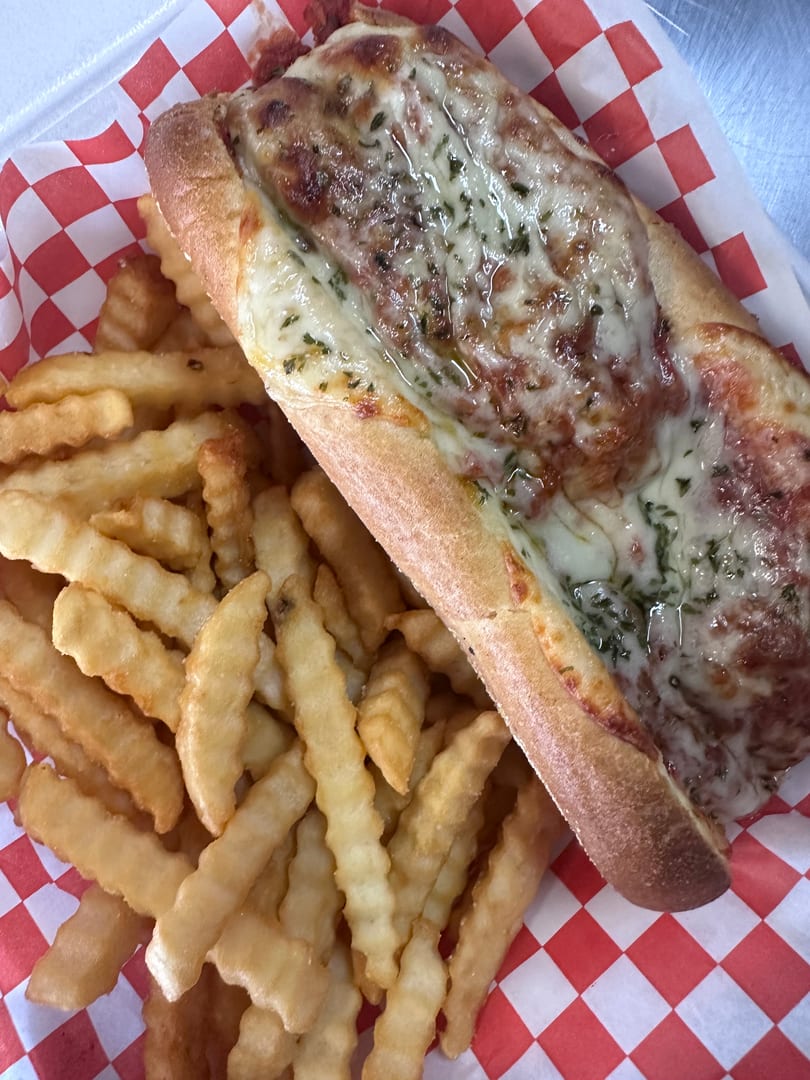 Meatball sub with melted cheese and herbs, served with crinkle-cut fries in a red and white checkered basket.