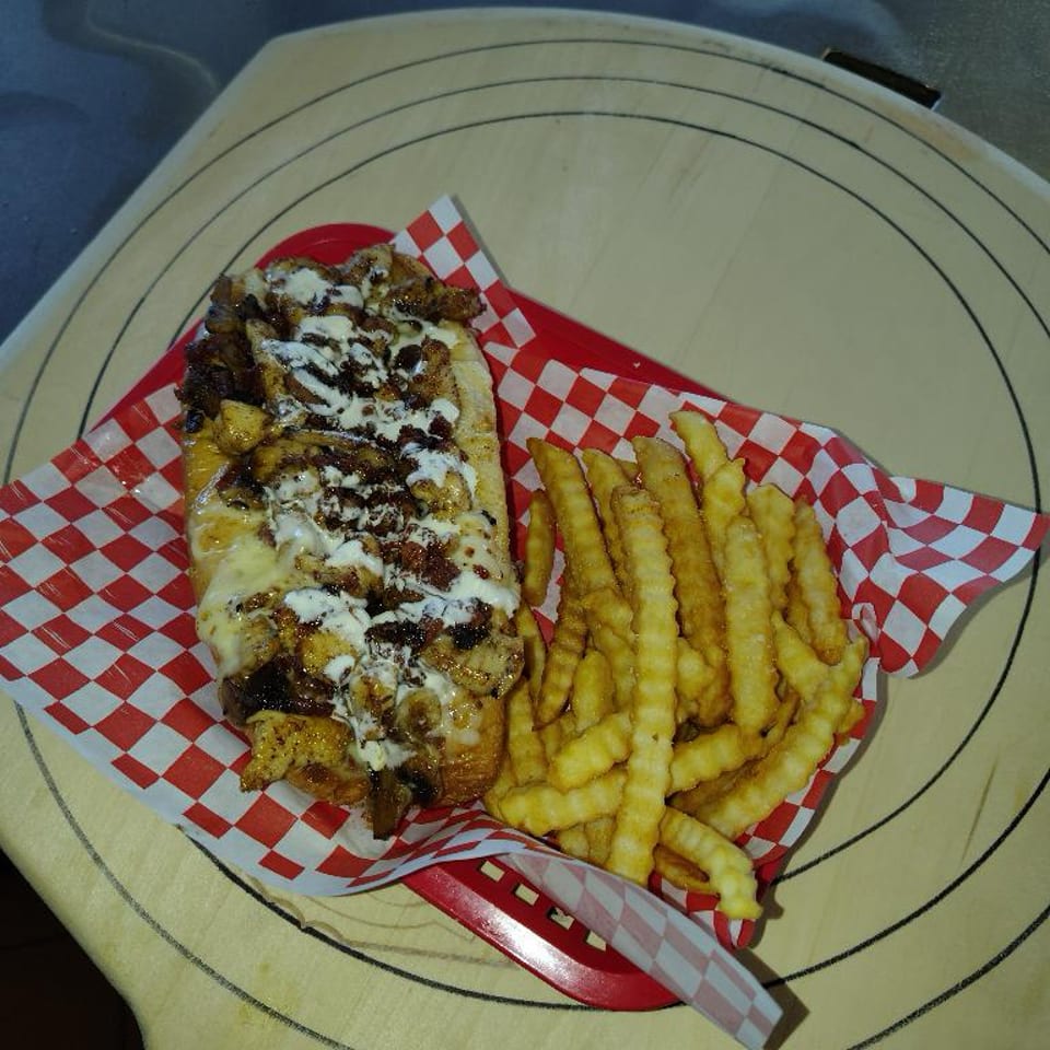 Cheesesteak sandwich with melted cheese and sauce, served with crinkle-cut fries in a red basket.