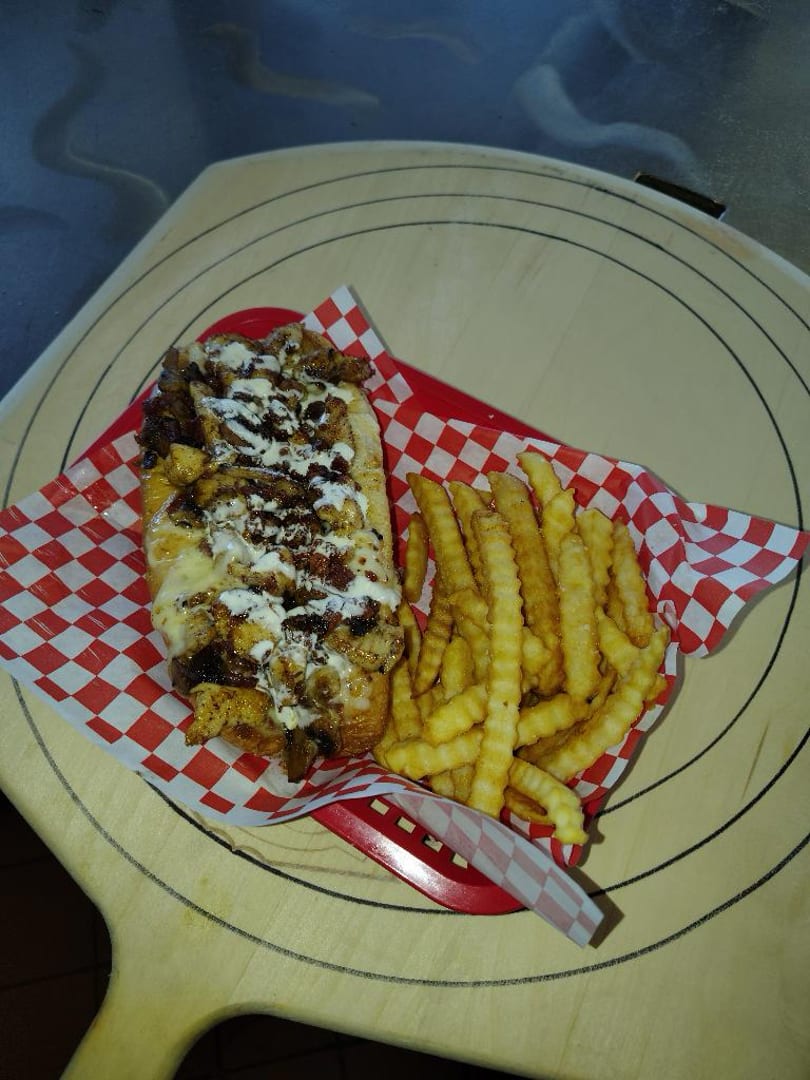 Cheesesteak sandwich with melted cheese and sauce, served with crinkle-cut fries in a red basket.